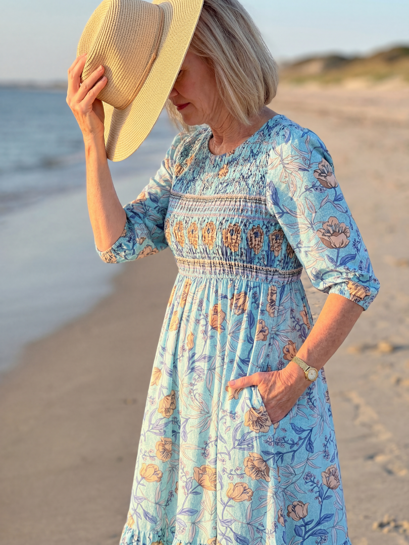Sky blue floral midi dress with gathered waist and three-quarter sleeves; straw hat held up