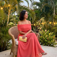 Off-shoulder draped midi dress in coral pink, seated side view with off-shoulder drape