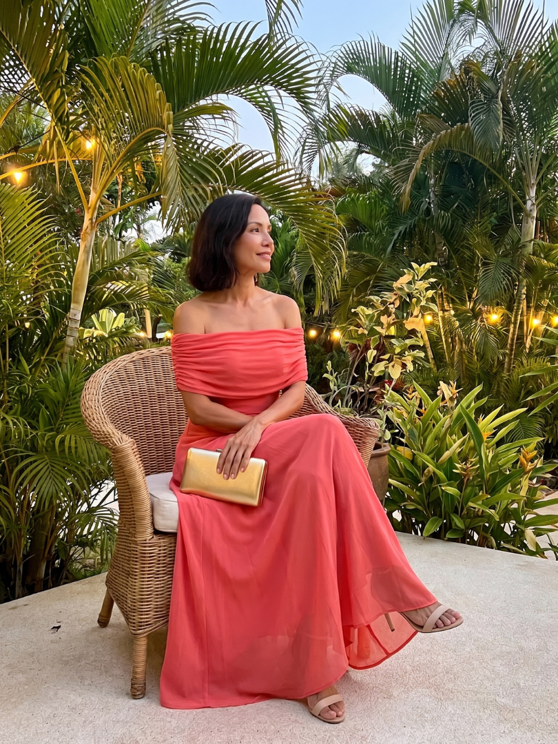 Off-shoulder draped midi dress in coral pink, seated side view with off-shoulder drape