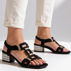 Black strappy sandals on feet with jeweled block heel and gold buckle details