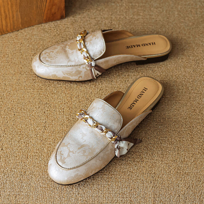 Pair of beige patterned leather mules with gold chain detail and bow at the strap