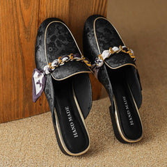 Pair of black patterned leather mules with gold chain across the upper and bow