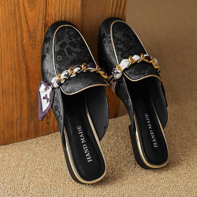 Pair of black patterned leather mules with gold chain across the upper and bow