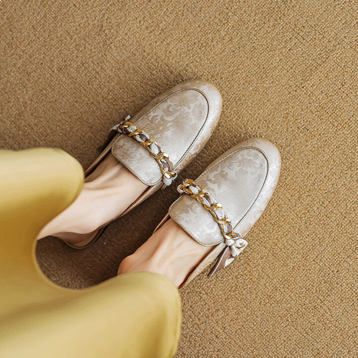 Beige patterned leather mules with gold chain across the upper and tied bow
