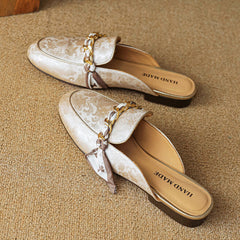 Beige patterned leather mules with gold chain trim and bow detail on the vamp