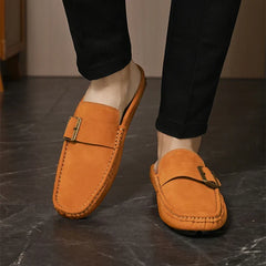 Tan suede mules with buckle strap on feet, stitched moc-toe design