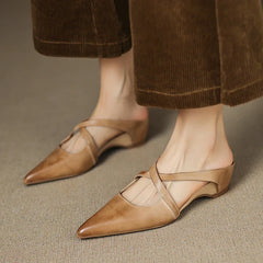 Tan leather mules on feet, pointed toe with cross slingback strap and low heel.