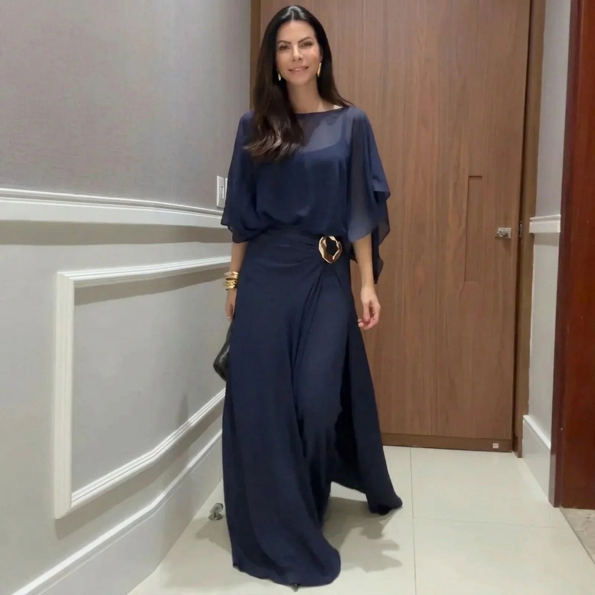 Navy long jumpsuit with sheer overlay cape sleeves and ring-buckle waist detail.