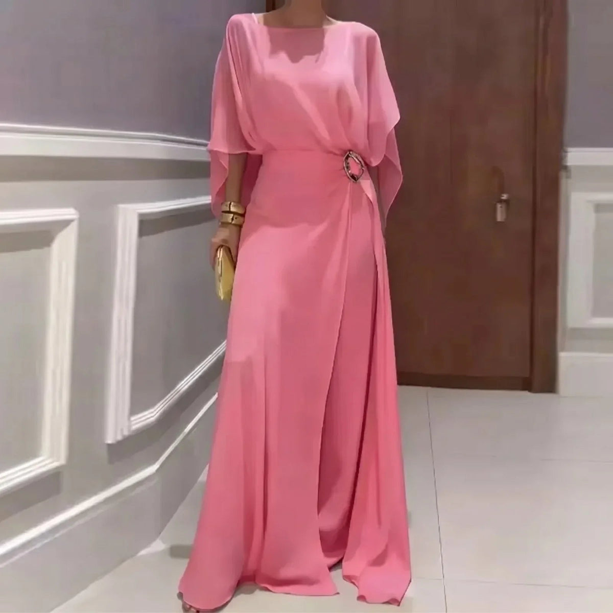 Pink long jumpsuit with sheer overlay cape sleeves and ring-buckle waist detail.